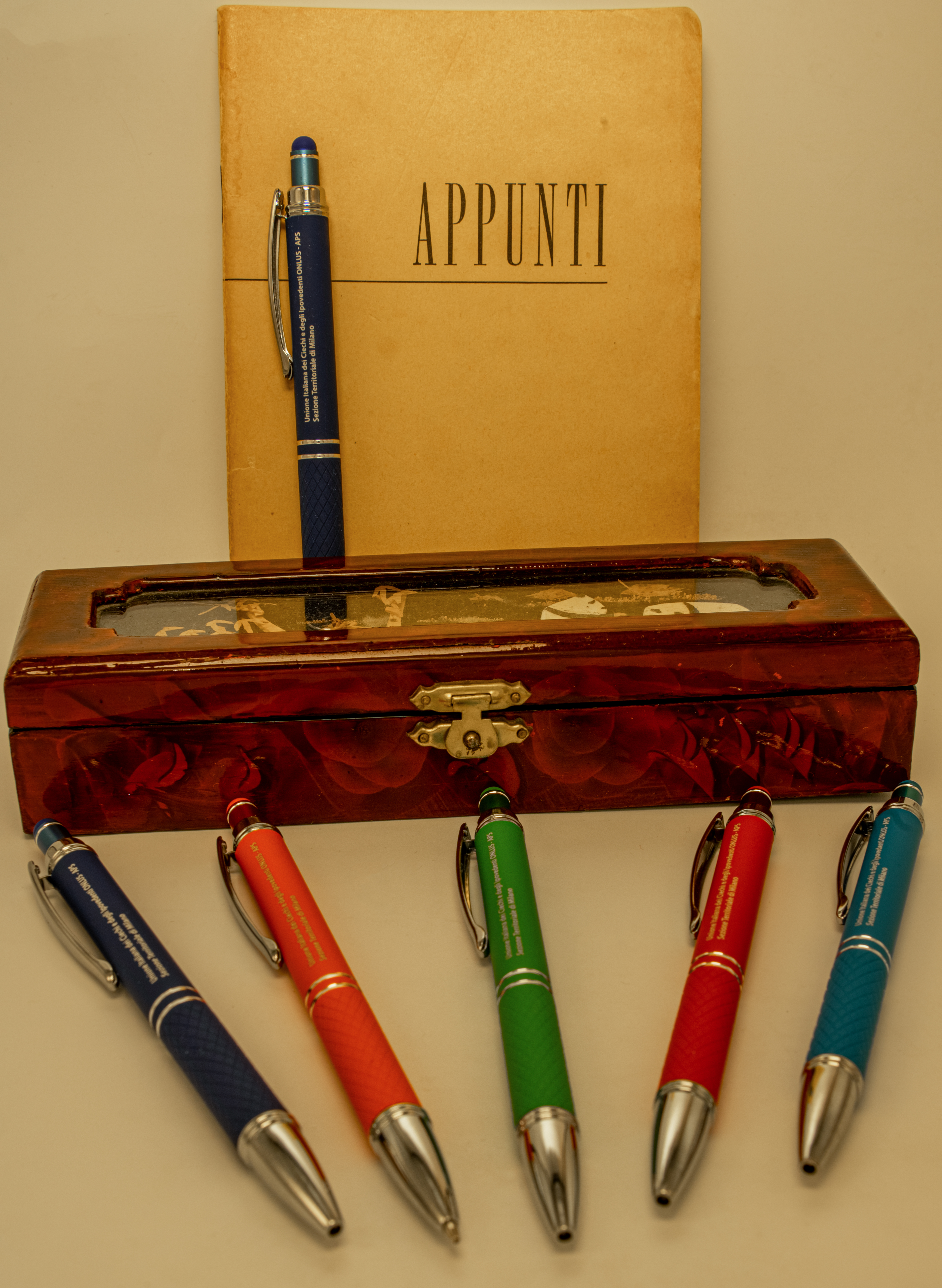 Le penne nelle cinque colorazioni e sullo sfondo un blocco per appunti ed un astuccio per penne d'epoca.